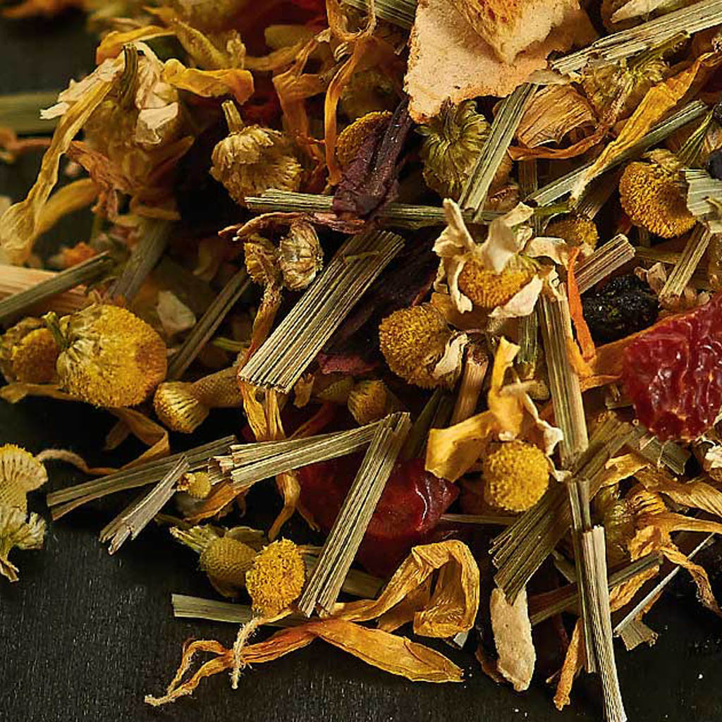 A close-up of Entspann Dich, a Kräuterteemischung featuring dried chamomile flowers, lemongrass, and botanicals on a dark surface—ideal for relaxing tea moments.